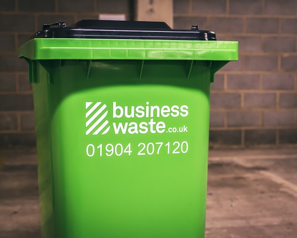 A green business waste bin is positioned against a brick wall. The bin is branded with a logo and contact information for a waste management company.