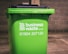 A green business waste bin is positioned against a brick wall. The bin is branded with a logo and contact information for a waste management company.