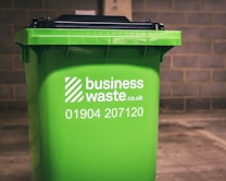 A green business waste bin is positioned against a brick wall. The bin is branded with a logo and contact information for a waste management company.