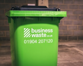 A green business waste bin is positioned against a brick wall. The bin is branded with a logo and contact information for a waste management company.