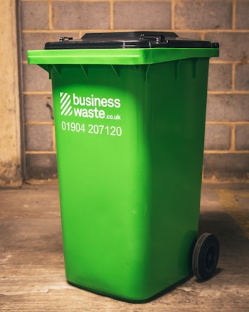 A bright green wheeled garbage bin with a black lid is positioned in front of a brick wall. The bin has the text 'business waste.co.uk 01904 207120' printed on its side in white.