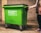 A person is pushing a large green waste bin with 'businesswaste.co.uk' and a phone number printed on the side. The bin has four wheels and is positioned in a dimly lit space with a wall of cinder blocks in the background.