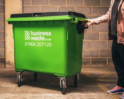 A person is pushing a large green waste bin with 'businesswaste.co.uk' and a phone number printed on the side. The bin has four wheels and is positioned in a dimly lit space with a wall of cinder blocks in the background.