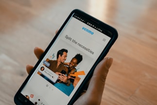 A person holds a smartphone displaying an app with a photo of two people smiling and using a phone together. Text on the screen reads 'Split the necessities'. Various app interface elements are visible, including icons and text for utilities and payment amounts.