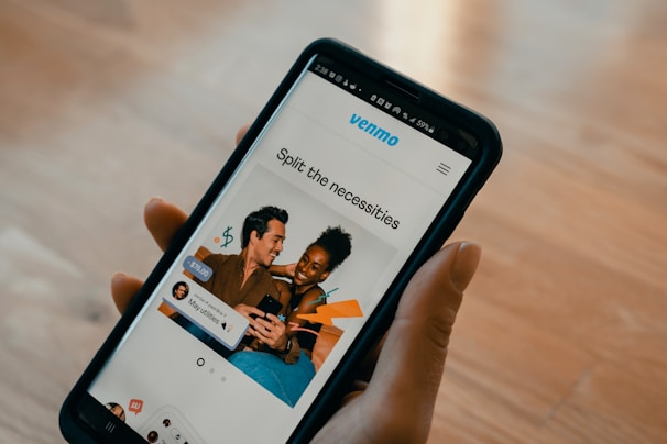 A person holds a smartphone displaying an app with a photo of two people smiling and using a phone together. Text on the screen reads 'Split the necessities'. Various app interface elements are visible, including icons and text for utilities and payment amounts.