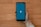 A hand is holding a smartphone displaying a blue screen with a white 'V' in the center. The phone is positioned over a light-colored wooden surface.