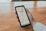 A person holding a smartphone displaying a financial app with a graph and balance information. The phone has a black case, and the interface shows details such as investment totals and changes. The background consists of a wooden table.