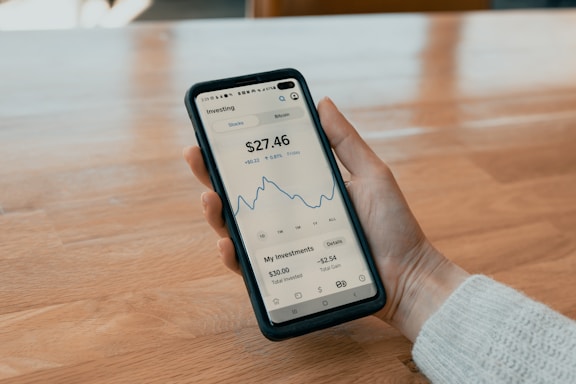 A person holding a smartphone displaying a financial app with a graph and balance information. The phone has a black case, and the interface shows details such as investment totals and changes. The background consists of a wooden table.