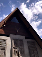 An A-frame wooden house with a triangular attic window and antlers mounted on the front. The structure features wooden shutters with intricate designs, complemented by the smooth, greyish plaster walls below. The sky is clear with a few scattered clouds, creating a peaceful atmosphere.