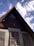 An A-frame wooden house with a triangular attic window and antlers mounted on the front. The structure features wooden shutters with intricate designs, complemented by the smooth, greyish plaster walls below. The sky is clear with a few scattered clouds, creating a peaceful atmosphere.