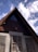An A-frame wooden house with a triangular attic window and antlers mounted on the front. The structure features wooden shutters with intricate designs, complemented by the smooth, greyish plaster walls below. The sky is clear with a few scattered clouds, creating a peaceful atmosphere.