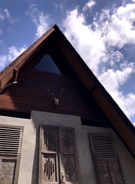 An A-frame wooden house with a triangular attic window and antlers mounted on the front. The structure features wooden shutters with intricate designs, complemented by the smooth, greyish plaster walls below. The sky is clear with a few scattered clouds, creating a peaceful atmosphere.