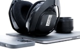 A pair of over-ear headphones with black padding and a sleek design rests on a closed laptop. Next to the laptop is a smartphone with a black case, and a camera lens is positioned slightly out of focus in the background.