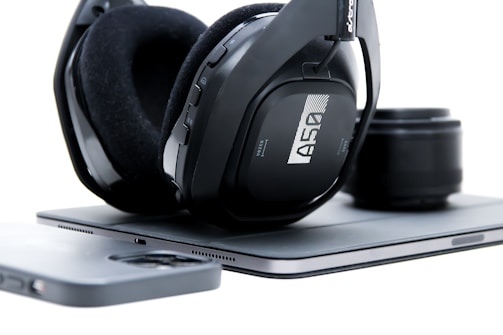 A pair of over-ear headphones with black padding and a sleek design rests on a closed laptop. Next to the laptop is a smartphone with a black case, and a camera lens is positioned slightly out of focus in the background.