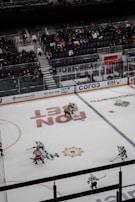 A hockey game is taking place on an indoor ice rink, with players in red and white jerseys actively engaged in play. The rink has advertisements on the ice and boards, and there's a moderate crowd in the stands, some wearing team colors.