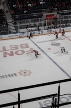 A professional ice hockey game taking place in an indoor arena with players from two teams, identifiable by red and white jerseys, actively engaged on the ice. The referee, wearing a black and white striped uniform, is monitoring the game. The audience is visible in the stands, which are partially filled.