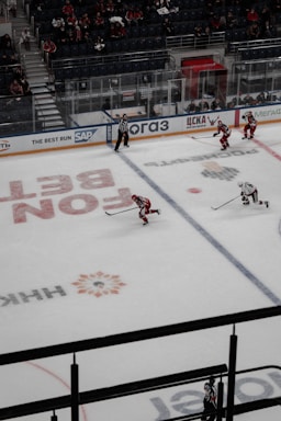 A professional ice hockey game taking place in an indoor arena with players from two teams, identifiable by red and white jerseys, actively engaged on the ice. The referee, wearing a black and white striped uniform, is monitoring the game. The audience is visible in the stands, which are partially filled.