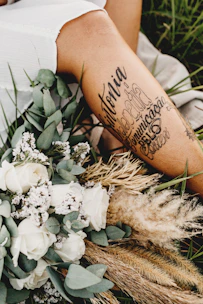 A close-up of a person's leg with artistic tattoos displayed prominently. The leg is surrounded by an arrangement of dried grass, white roses, and other small flowers. The setting appears to be outdoors, with green grass visible in the background, creating a serene and rustic atmosphere.
