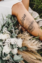 A close-up of a person's leg with artistic tattoos displayed prominently. The leg is surrounded by an arrangement of dried grass, white roses, and other small flowers. The setting appears to be outdoors, with green grass visible in the background, creating a serene and rustic atmosphere.