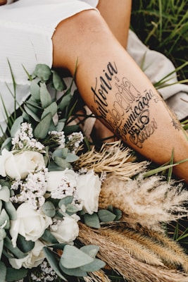 A close-up of a person's leg with artistic tattoos displayed prominently. The leg is surrounded by an arrangement of dried grass, white roses, and other small flowers. The setting appears to be outdoors, with green grass visible in the background, creating a serene and rustic atmosphere.