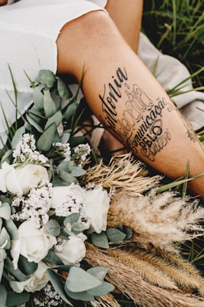 A close-up of a person's leg with artistic tattoos displayed prominently. The leg is surrounded by an arrangement of dried grass, white roses, and other small flowers. The setting appears to be outdoors, with green grass visible in the background, creating a serene and rustic atmosphere.