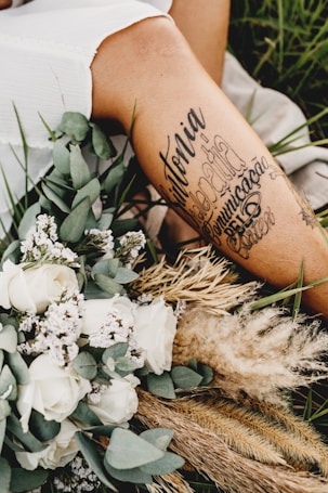 A close-up of a person's leg with artistic tattoos displayed prominently. The leg is surrounded by an arrangement of dried grass, white roses, and other small flowers. The setting appears to be outdoors, with green grass visible in the background, creating a serene and rustic atmosphere.