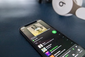 A smartphone displays a Spotify playlist interface with a song playing, showing various controls and song details. Beside it, white and brown headphones with a distinctive logo are partially visible on a dark surface.
