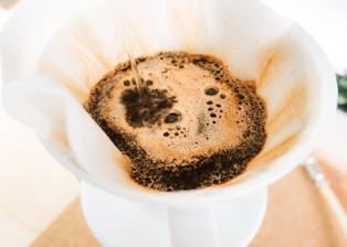 A close-up view of coffee being brewed using a pour-over method. The coffee is filtering through a white paper filter and forming a frothy, bubbly liquid with dark grounds visible.