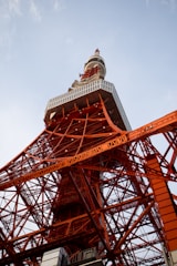 A large metal tower structure with a lattice framework, painted in a bright orange-red color, stands against a clear blue sky. The tower is intricately constructed with overlapping beams and various geometric shapes, showcasing an impressive engineering design.