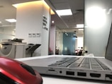 A modern office environment with a laptop on a white desk in the foreground. A red computer mouse is also visible. In the background, there is an office printer and a wall with motivational text. The office space is well-lit with natural and artificial light, and there are glass partitions.