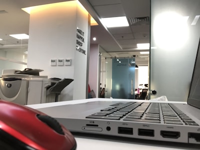 A modern office environment with a laptop on a white desk in the foreground. A red computer mouse is also visible. In the background, there is an office printer and a wall with motivational text. The office space is well-lit with natural and artificial light, and there are glass partitions.