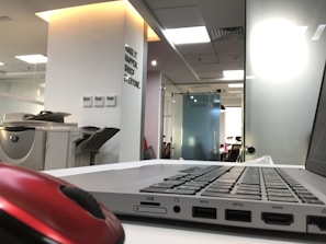 A modern office environment with a laptop on a white desk in the foreground. A red computer mouse is also visible. In the background, there is an office printer and a wall with motivational text. The office space is well-lit with natural and artificial light, and there are glass partitions.