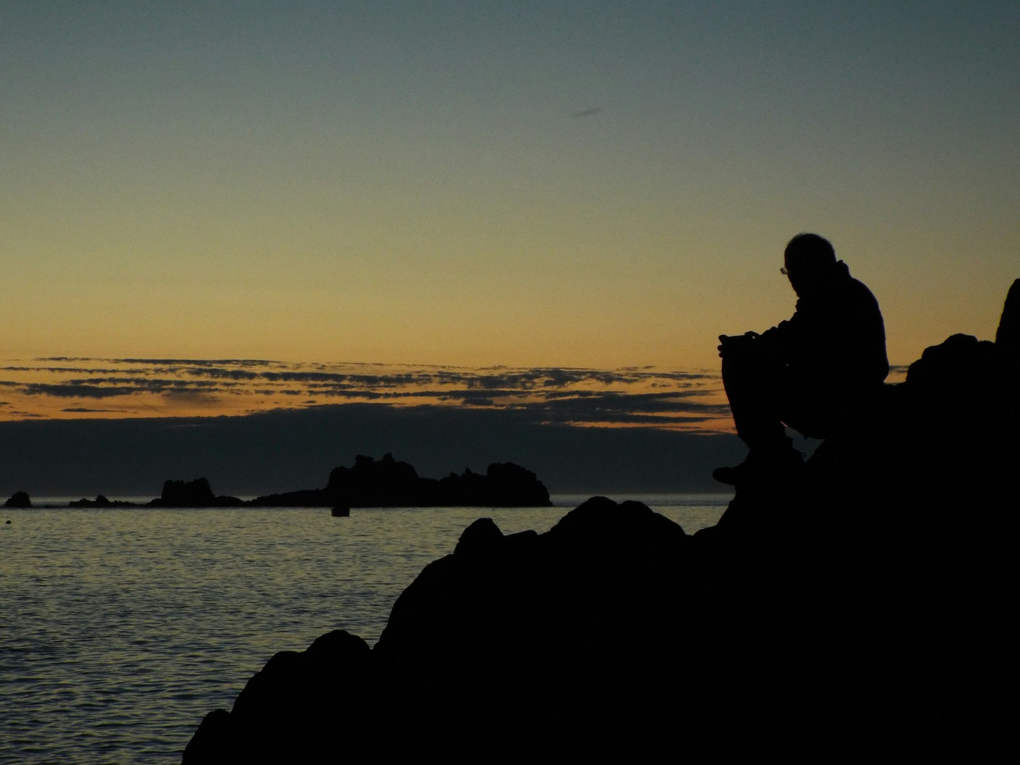 A silhouette of a man sitting on a rock looking out at a golden sunset sea sky.