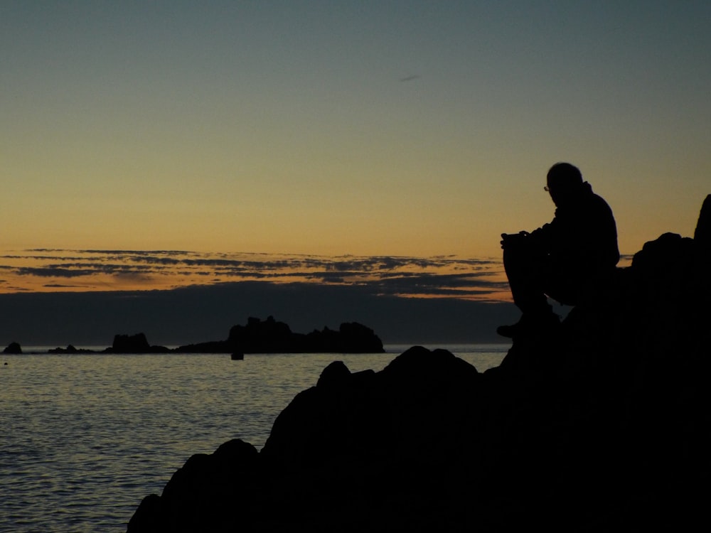 silhouette of man and woman sitting on rock near body of water during sunset