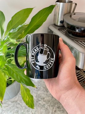 A hand holds a black coffee mug with a printed design featuring a coffee pot and the words 'Pot Head Coffee'. In the background, there is a green potted plant and a stainless steel coffee machine with a glass pot on top. The setting appears to be a kitchen.