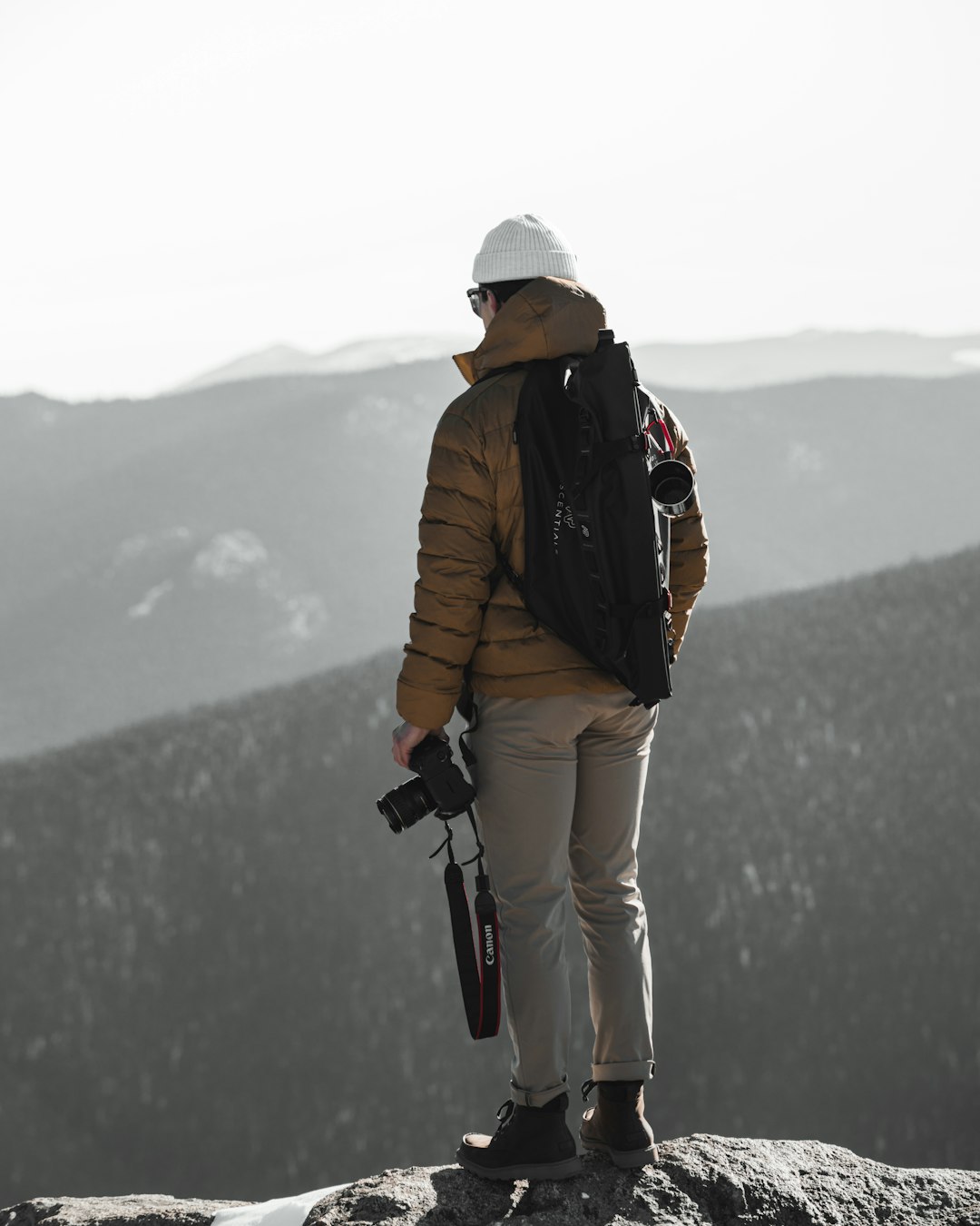 man in brown jacket and white pants with black backpack standing on rock during daytime