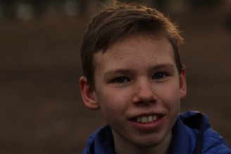 A young person with short hair and a blue jacket is smiling outdoors. The background is blurred, giving focus to the individual's expression and features.