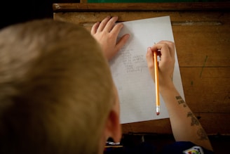 A person is writing on lined paper with a pencil, listing names of states such as Washington, Kansas, and Kentucky. The setting appears to be a wooden desk, and the person has short blond hair and tattoos on their arm.