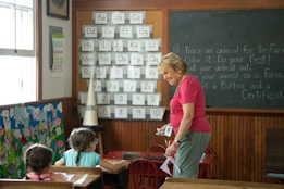 A classroom setting with children sitting at desks and an adult woman standing and interacting with them. The classroom has posters and decorations on the walls, including an alphabet and numbers chart, and a chalkboard with instructions. The woman is holding some items in her hands and seems to be engaging with the children, who are looking at her attentively.