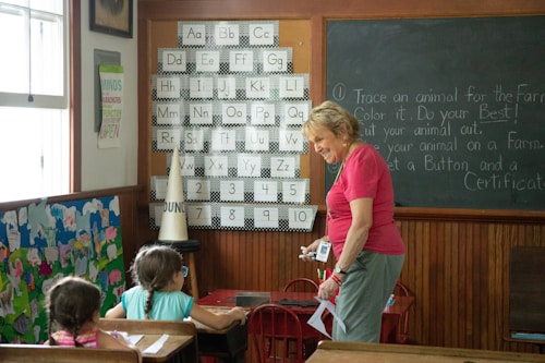 A classroom setting with children sitting at desks and an adult woman standing and interacting with them. The classroom has posters and decorations on the walls, including an alphabet and numbers chart, and a chalkboard with instructions. The woman is holding some items in her hands and seems to be engaging with the children, who are looking at her attentively.