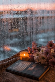 A cozy scene featuring a lit candle labeled 'rosemary sage' next to a stack of flowers and a book on a textured fabric surface. Raindrops are visible on a window with a blurred cityscape and sunset in the background, creating a warm and serene atmosphere.