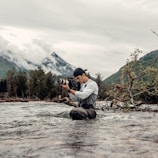A person is standing knee-deep in a flowing river, holding a professional camera. The background features a mountainous landscape with misty clouds, green trees, and foliage. The person appears to be wearing a cap and waders, suggesting they are engaged in outdoor photography.