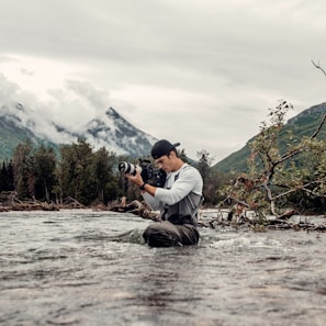 A person is standing knee-deep in a flowing river, holding a professional camera. The background features a mountainous landscape with misty clouds, green trees, and foliage. The person appears to be wearing a cap and waders, suggesting they are engaged in outdoor photography.