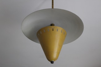 A vintage ceiling lamp with a unique conical design. The lamp has a mustard yellow shade with small perforations near the top, hanging from the ceiling by a central rod. The bottom of the cone features a small decorative element.