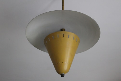 A vintage ceiling lamp with a unique conical design. The lamp has a mustard yellow shade with small perforations near the top, hanging from the ceiling by a central rod. The bottom of the cone features a small decorative element.