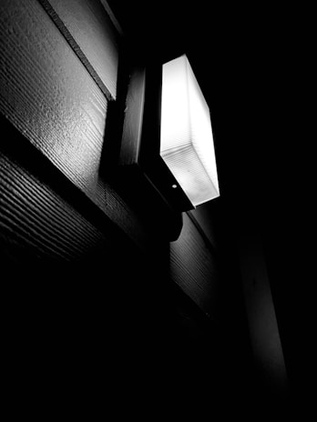 A black and white image featuring an illuminated outdoor wall light fixture mounted on a wooden-paneled wall. The light casts sharp shadows and contrasts against the dark surroundings, highlighting the texture of the wall.