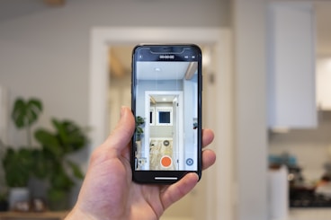 A hand holds a smartphone in a modern, well-lit room. The phone's screen displays a live camera view showing an interior hallway with a plant and bright lighting. In the background, a blurred view of a kitchen is visible with cabinets and appliances.