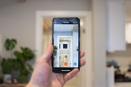 A hand holds a smartphone in a modern, well-lit room. The phone's screen displays a live camera view showing an interior hallway with a plant and bright lighting. In the background, a blurred view of a kitchen is visible with cabinets and appliances.