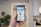 A hand holds a smartphone in a modern, well-lit room. The phone's screen displays a live camera view showing an interior hallway with a plant and bright lighting. In the background, a blurred view of a kitchen is visible with cabinets and appliances.