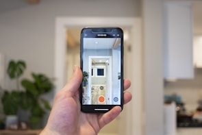 A hand holds a smartphone in a modern, well-lit room. The phone's screen displays a live camera view showing an interior hallway with a plant and bright lighting. In the background, a blurred view of a kitchen is visible with cabinets and appliances.